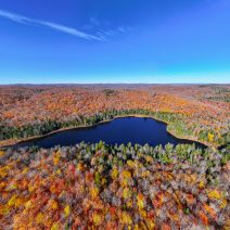 Photographie aérienne en plongée montrant un lac aux eaux bleues profondes niché au cœur d'une forêt laurentienne en automne. Les arbres arborent des couleurs vives de saison allant du rouge, orange et jaune au vert des conifères, sous un grand ciel bleu dégagé.